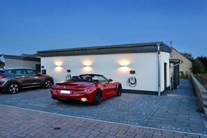 Ein modernes Gebäude mit weißer Fassade, davor ein roter Cabrio und ein brauner SUV zur Linke.