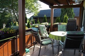 Eine überdachte Terrasse mit rundem Tisch und Stühlen, umgeben von Pflanzen und einem Holzzaun.