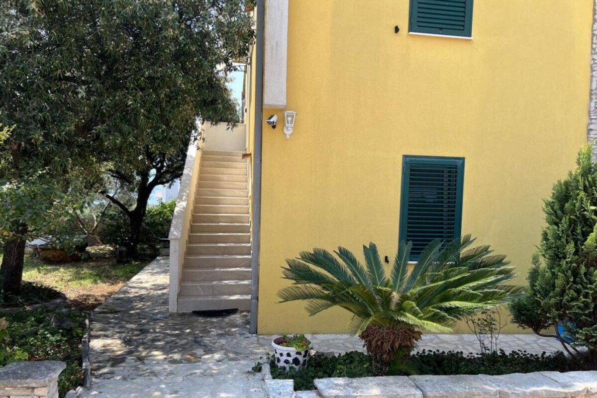 A yellow building with a staircase leading to a door, surrounded by greenery and a stone pathway.