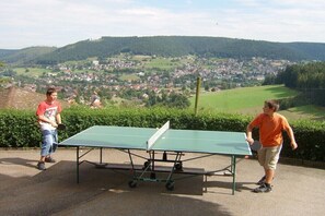 Zwei Personen spielen Tischtennis im Freien, mit einer malerischen Dorflandschaft im Hintergrund.