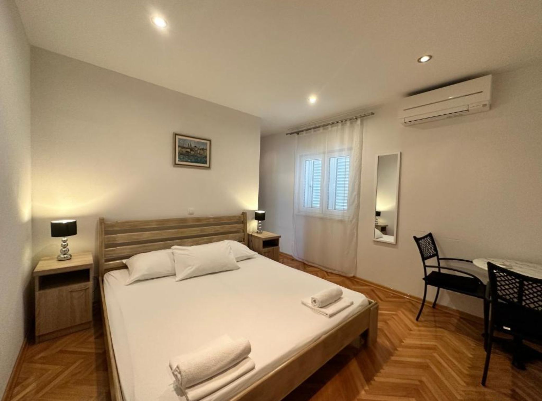 A hotel room with a bed, bedside lamps, a desk with a chair, a window with curtains, an air conditioning unit, and a painting on the wall.