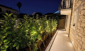 A pathway lined with illuminated plants leading to a stone building with a balcony.