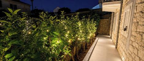 A pathway lined with illuminated plants leading to a stone building with a balcony.