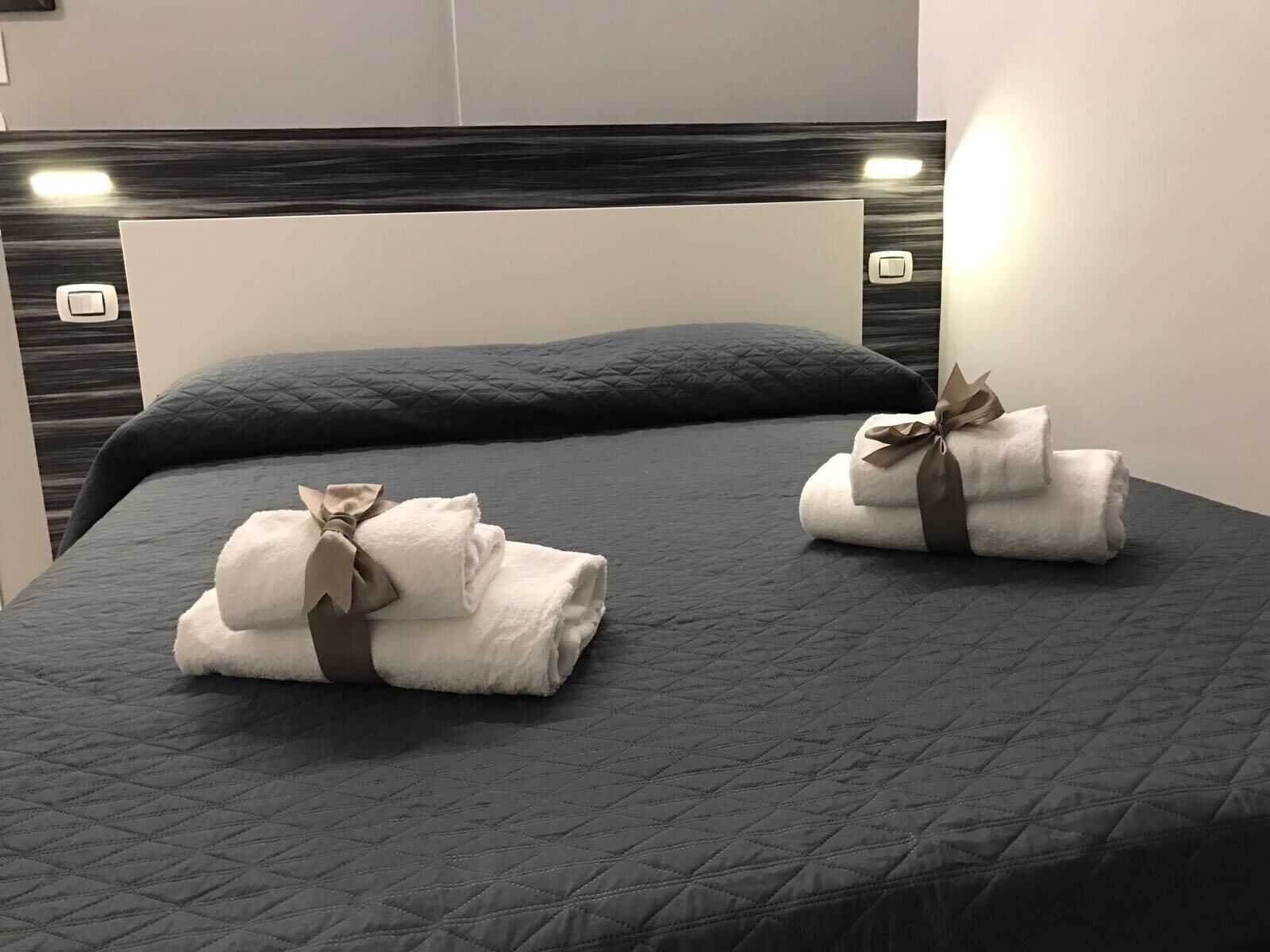 Un lit recouvert d’une courtepointe grise à motifs, deux serviettes soigneusement pliées, ornées de rubans marron, et une tête de lit dotée d’un interrupteur.