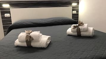 Een bed met een geborduurde grijze dekbedovertrek, twee keurig opgevouwen handdoeken met bruine linten, en een hoofdbord met een lichtschakelaar.
