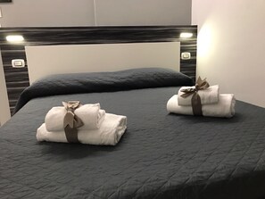A bed with a quilted grey bedspread, two neatly folded towels with brown ribbons, and a headboard with a light switch. - Standard Room 1 2 people in Piazza San Giovanni (ROMA)