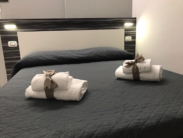 Una cama con colcha gris acolchada, dos toallas dobladas con cintas marrones y un cabecero con interruptor de luz.