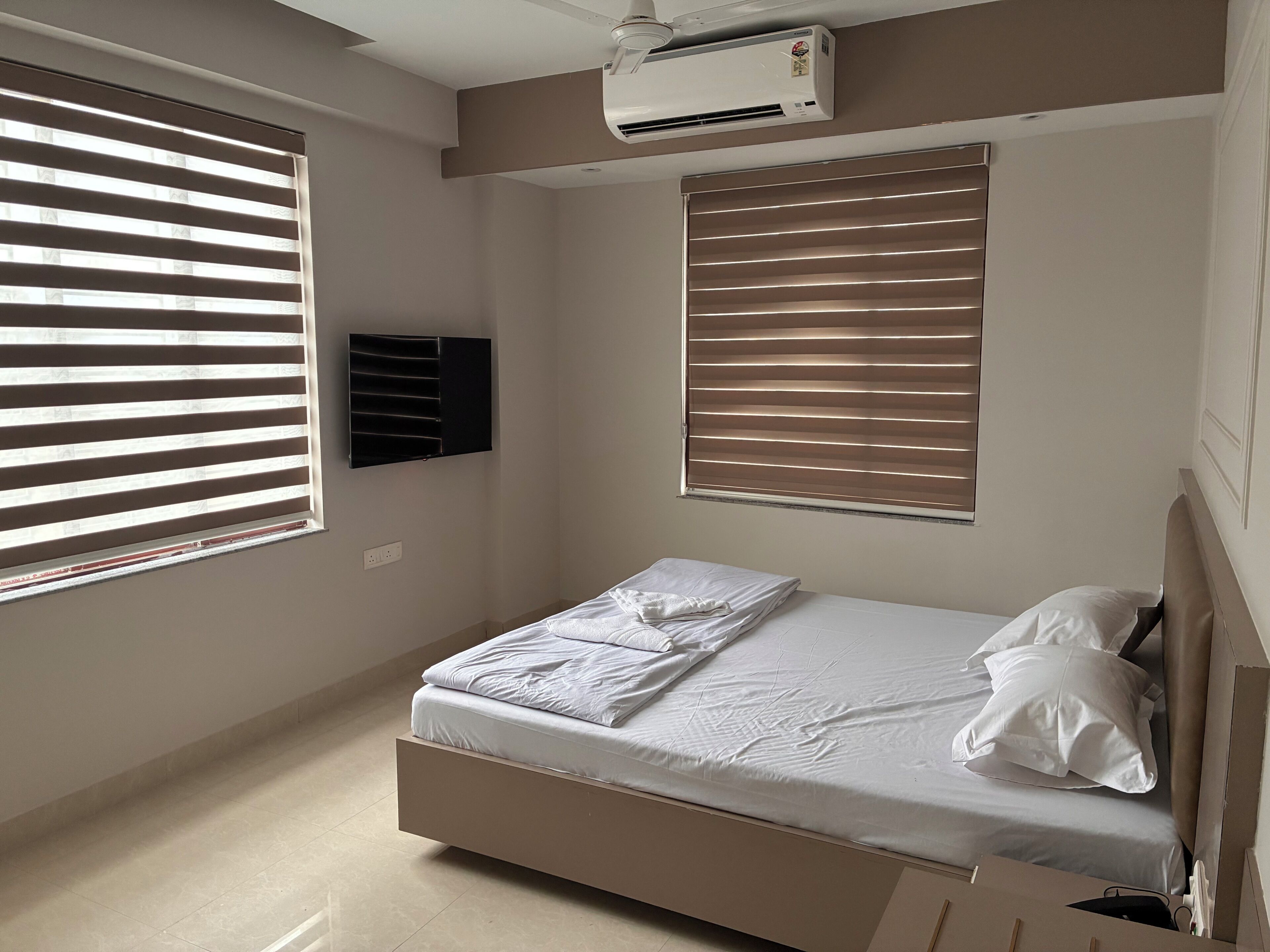 A bedroom with a bed, two windows with blinds, an air conditioning unit, and a wall-mounted TV.