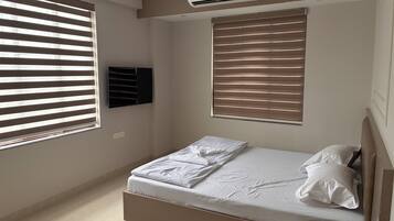 Un dormitorio con cama, dos ventanas con persianas, un equipo de aire acondicionado y un televisor montado en la pared.