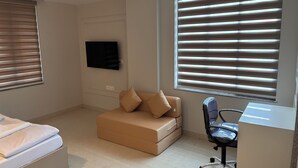 Un dormitorio con cama, sofá, escritorio y ventanas grandes con persianas.