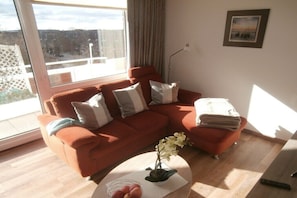 A living room with a red sofa, a coffee table with a plant and fruit bowl, a remote control, and a window with a view of a fence and trees. - 348 in the Trafalgar House (Cuxhaven - Döse)