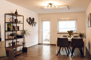 Una sala de estar moderna con comedor, que incluye piso de madera, una estantería, una mesa de centro y una mesa de comedor preparada para dos personas.