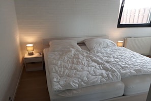 Ein Einzelbett mit weißer Decke, zwei Nachttischlampen, eine weiße Ziegelwand und ein Fenster mit Jalousien.