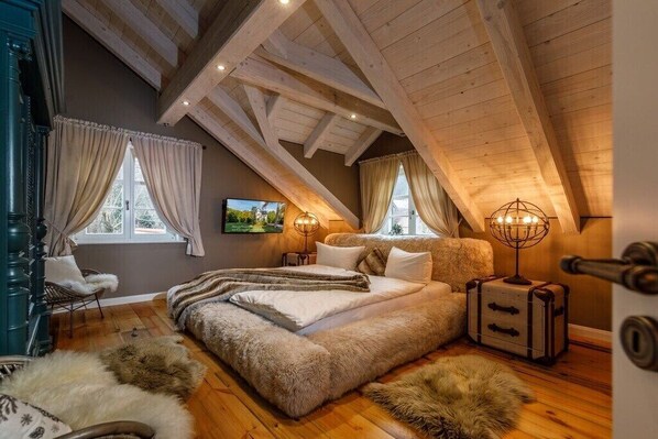 A bedroom with a sloped ceiling, wooden beams, a bed with a fluffy blanket, a lamp, and a suitcase.