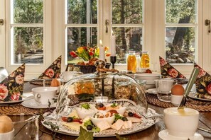 A breakfast setting with a glass dome covering a selection of cheeses, fruits, and bread, with a view of trees through the windows.