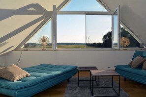 Ein Zimmer mit schrägen Wänden, einem großen Fenster, zwei blauen Sofas, einem kleinen Tisch und Blick auf ein Feld und einen Windm{"u}{"u}lle.