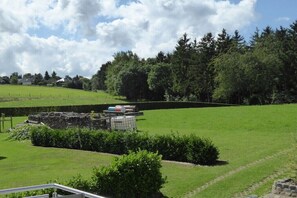 Ein grasbewachsenes Feld mit einer Steinstruktur, vermutlich eine historische Stätte, umgeben von Bäumen und blauem Himmel.