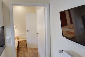 Ein Zimmer mit einem Bett, einem Nachttisch, einem an der Wand befestigten Fernseher und einer Tür zu einem anderen Raum.