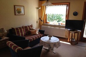 Ein Wohnzimmer mit einer Couch, einem kleinen Tisch, einem Fernseher und einem Fenster mit Blick auf Grünflächen.