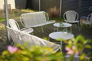 A patio with white metal chairs and tables, surrounded by plants and a brick wall.