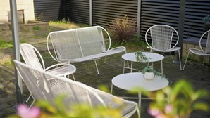 Eine Terrasse mit weißen Metallstühlen und -tischen, umgeben von Grünpflanzen und einem Pflasterboden.