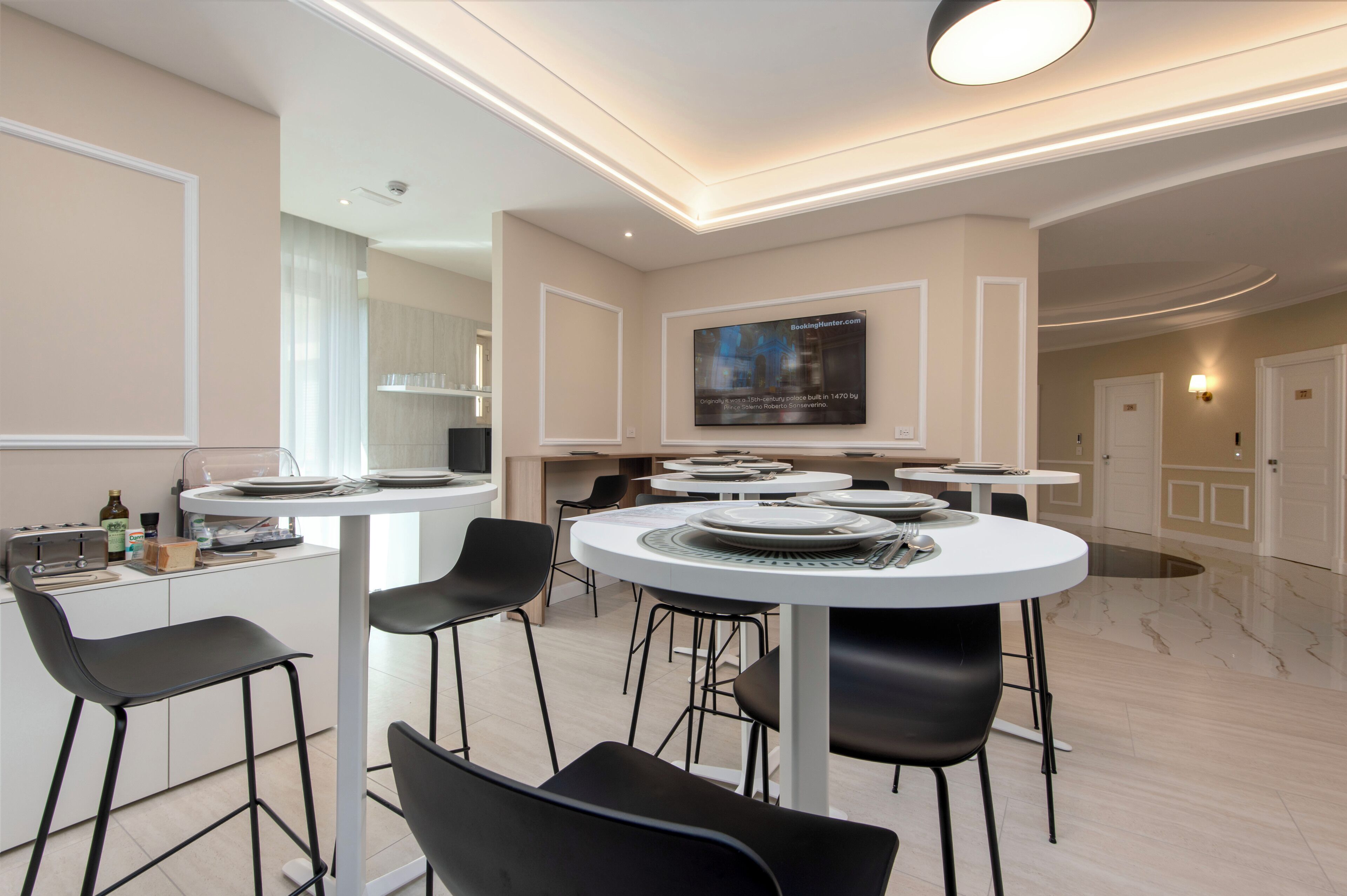 A modern kitchen with white cabinets, black bar stools, and a large flat-screen TV mounted on the wall.