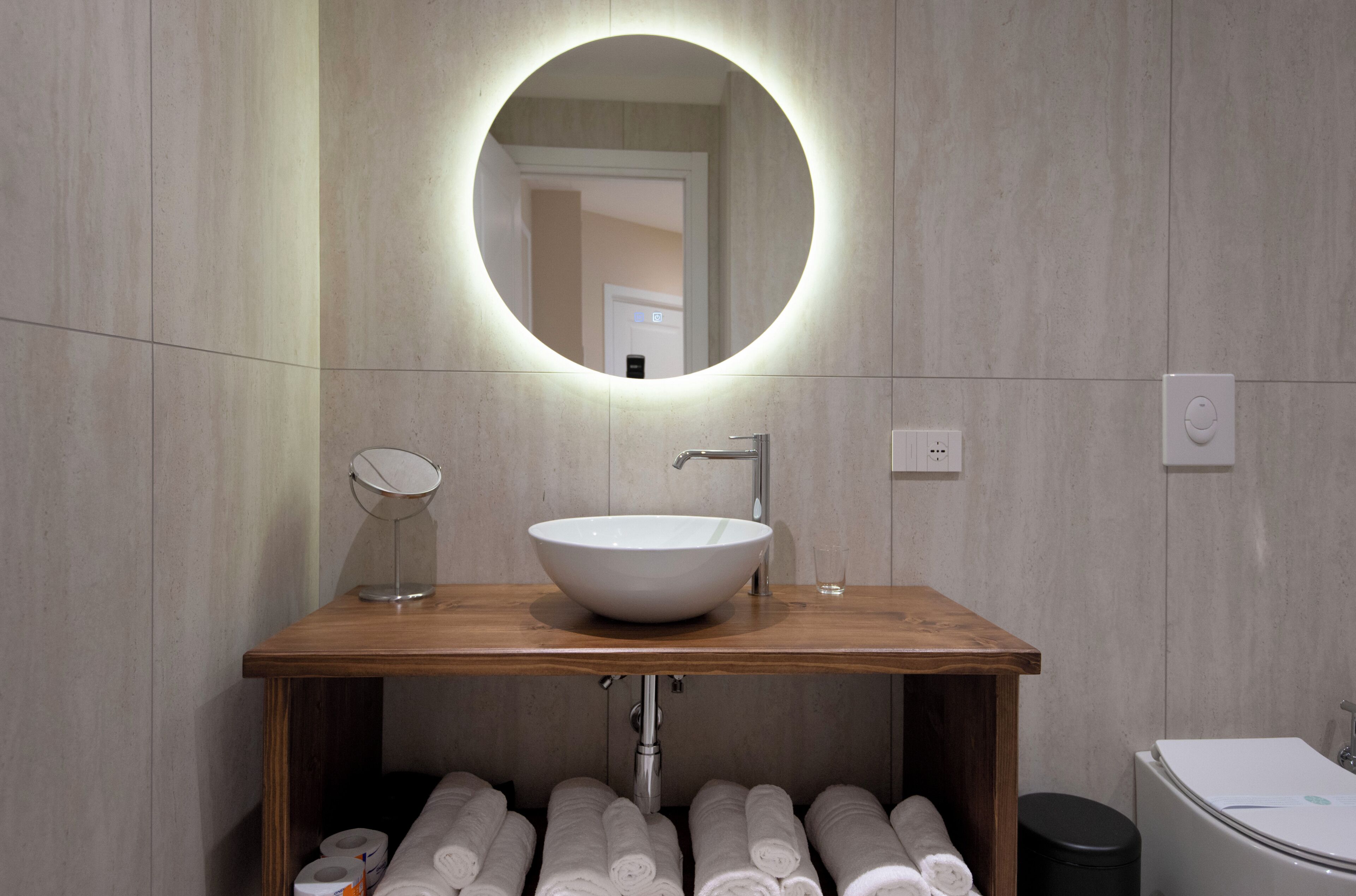 A modern bathroom with a wooden vanity, a round mirror, a white sink, and neatly rolled towels.