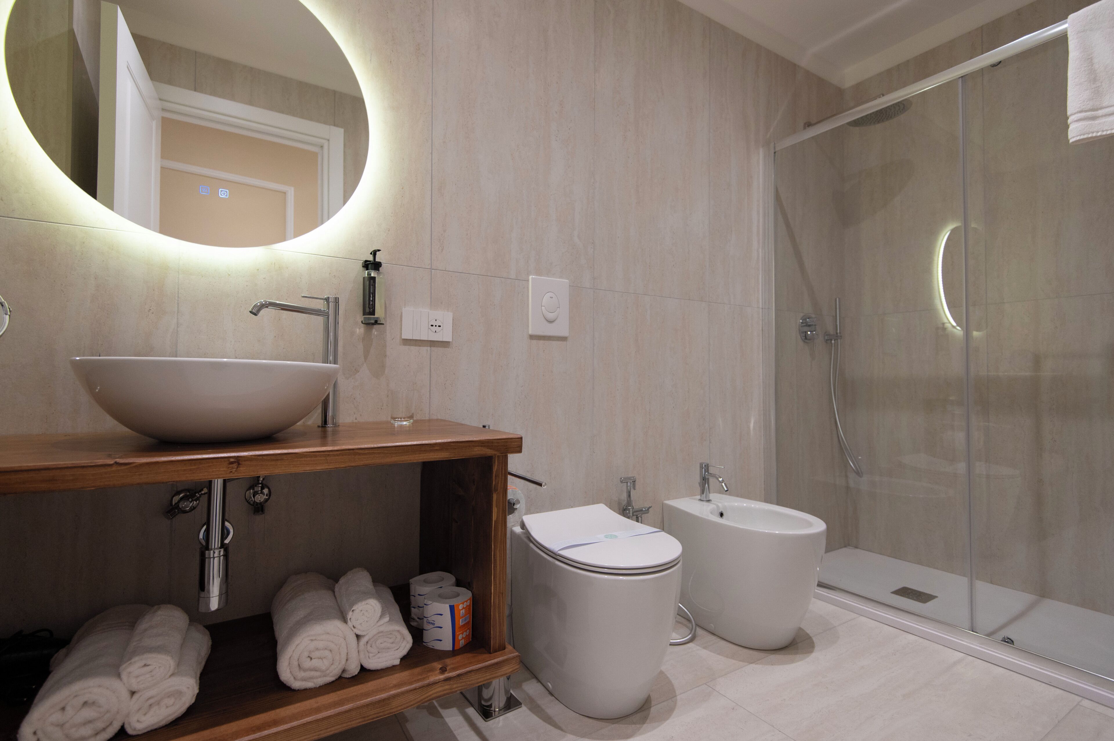 Une salle de bain moderne avec un grand miroir ovale, un lavabo sur colonne, des toilettes, un bidet et une douche à parois de verre.
