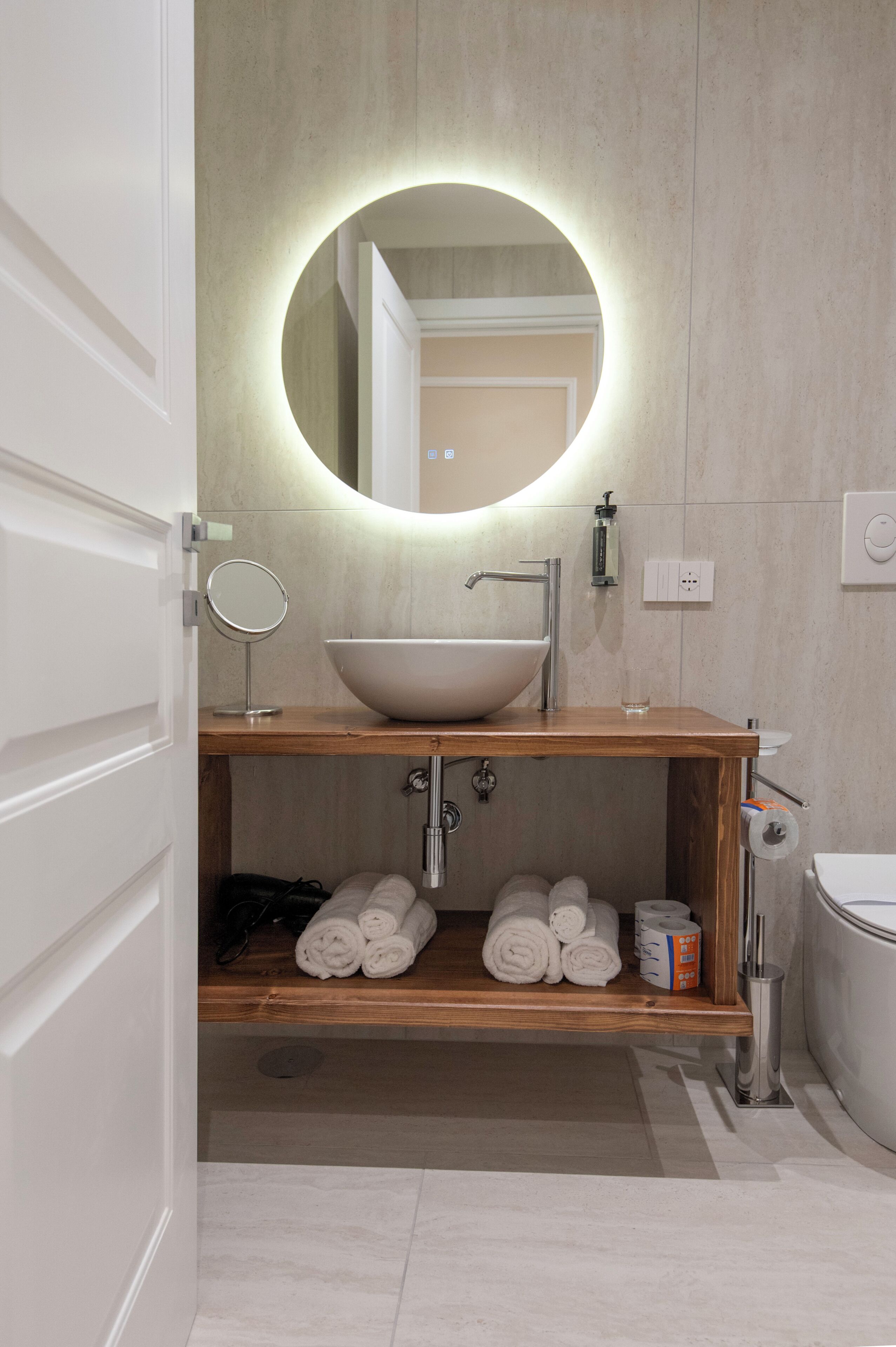 A modern bathroom with a wooden vanity, a round mirror, a vessel sink, and neatly rolled towels on a shelf.