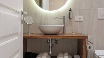 A modern bathroom with a wooden vanity, a round mirror, a vessel sink, and neatly rolled towels on a shelf.