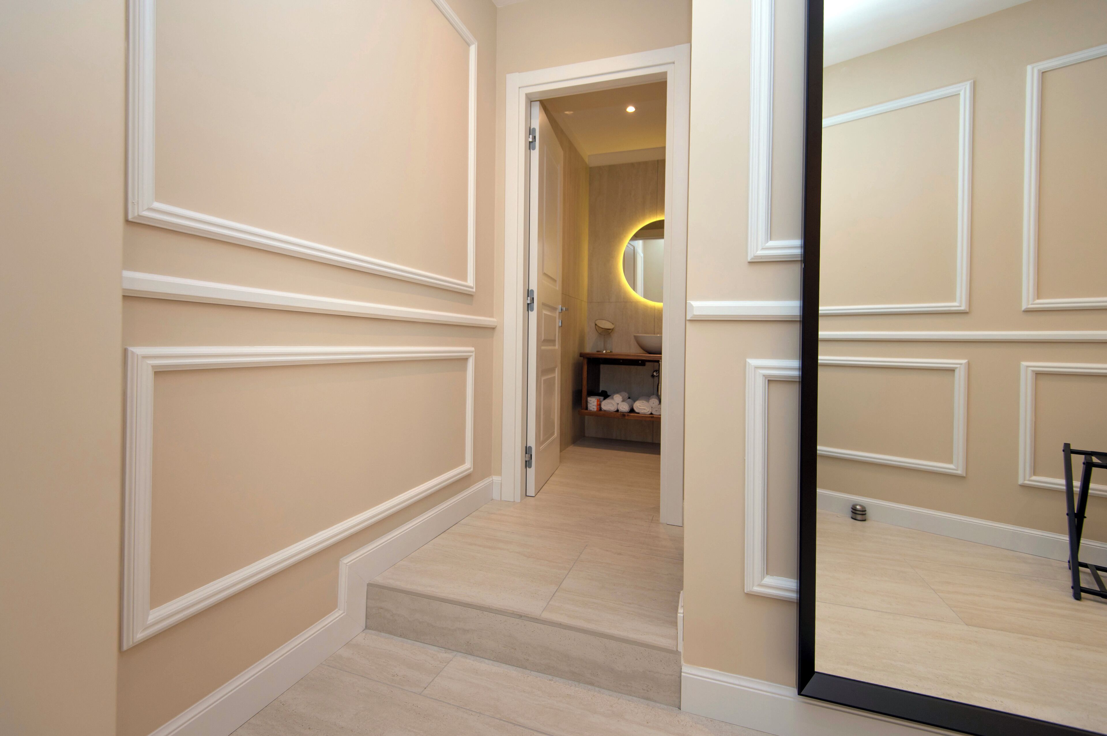 Un pasillo con pared de color claro, suelo de madera y una puerta que da a una habitación con un espejo y una repisa.