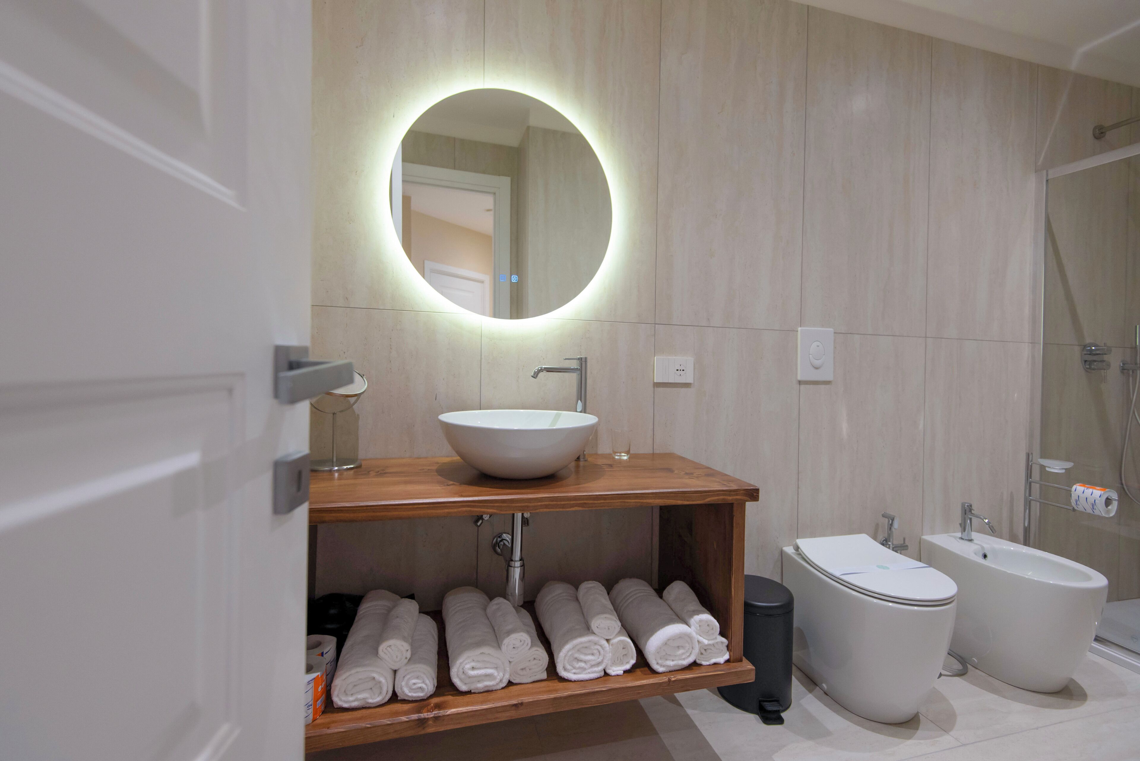 A modern bathroom with a round mirror, a sink, a bidet, and a bathtub.