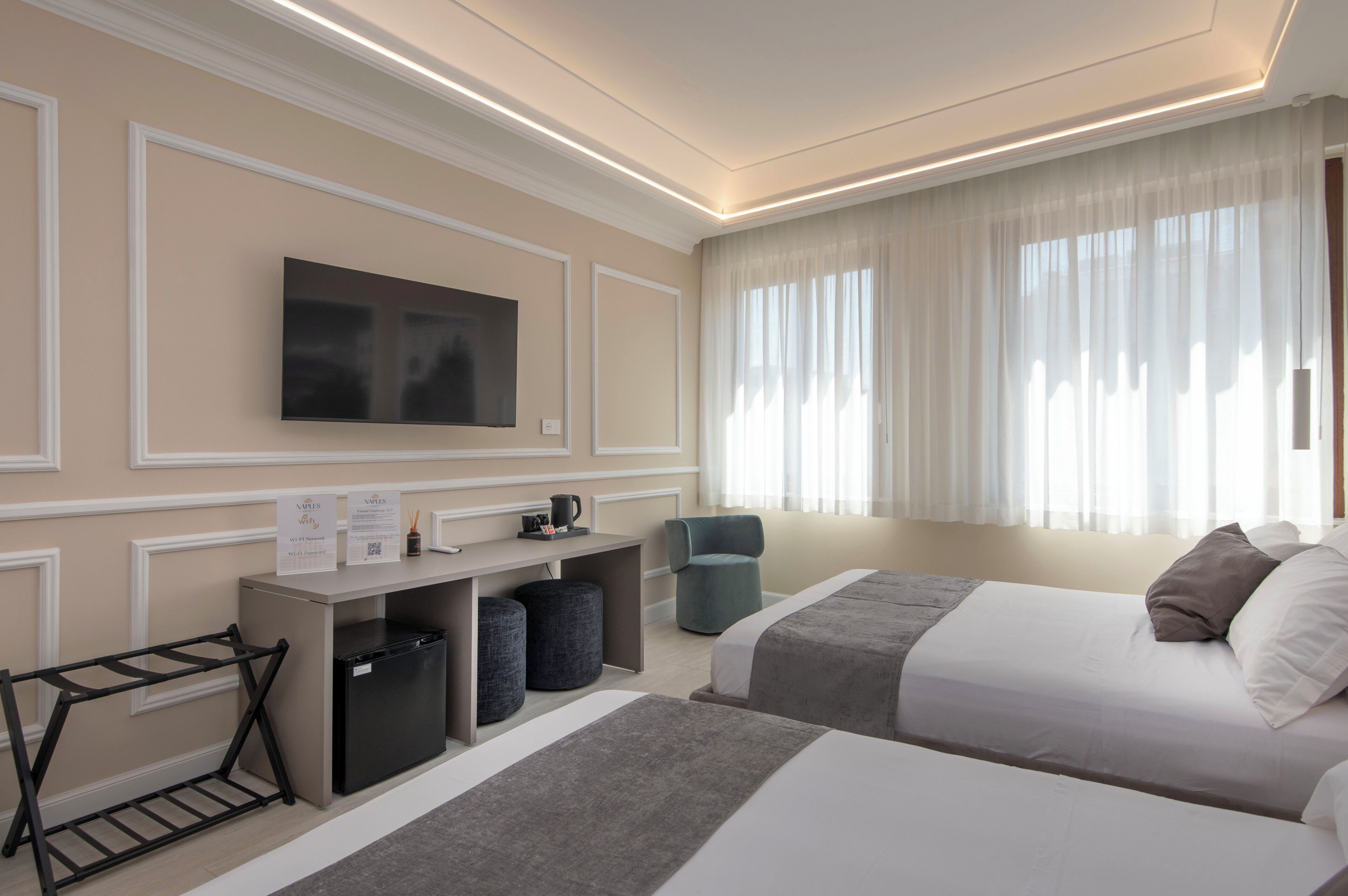 A hotel room with two beds, a TV, a desk with a computer, and a window with sheer curtains.