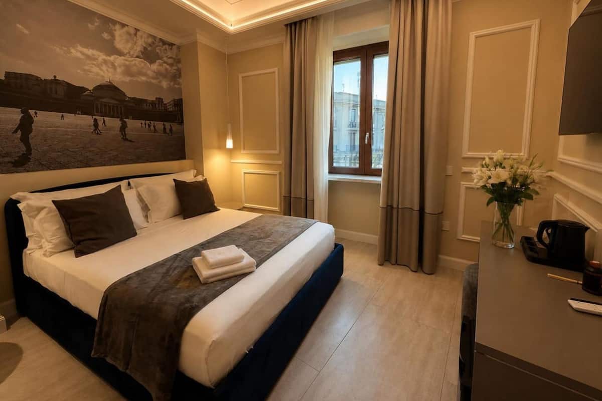A hotel room with a large bed, a painting on the wall, a window with curtains, and a desk with a coffee maker.