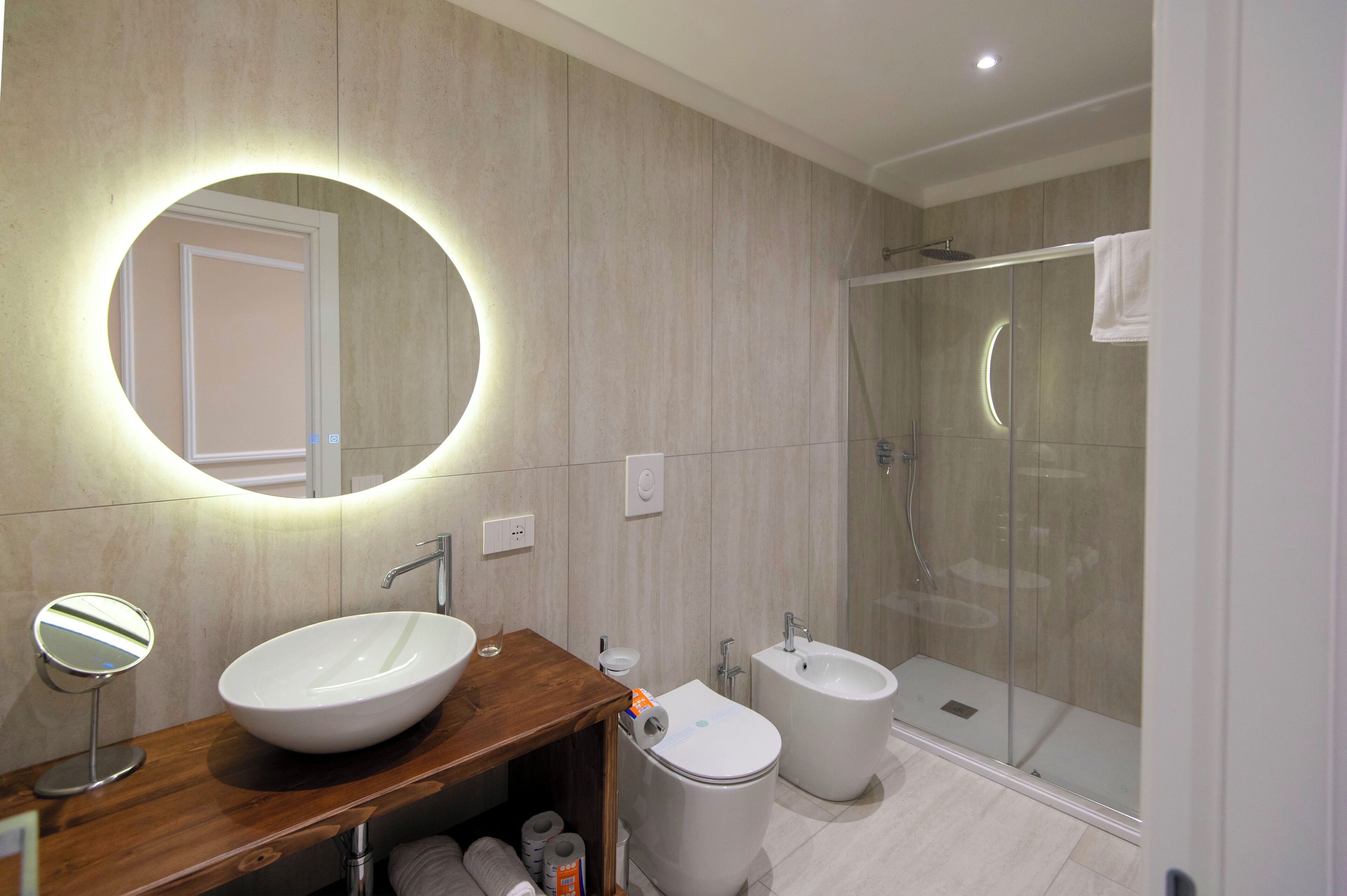A modern bathroom with a wooden vanity, a large mirror, a bidet, and a glass-enclosed shower.
