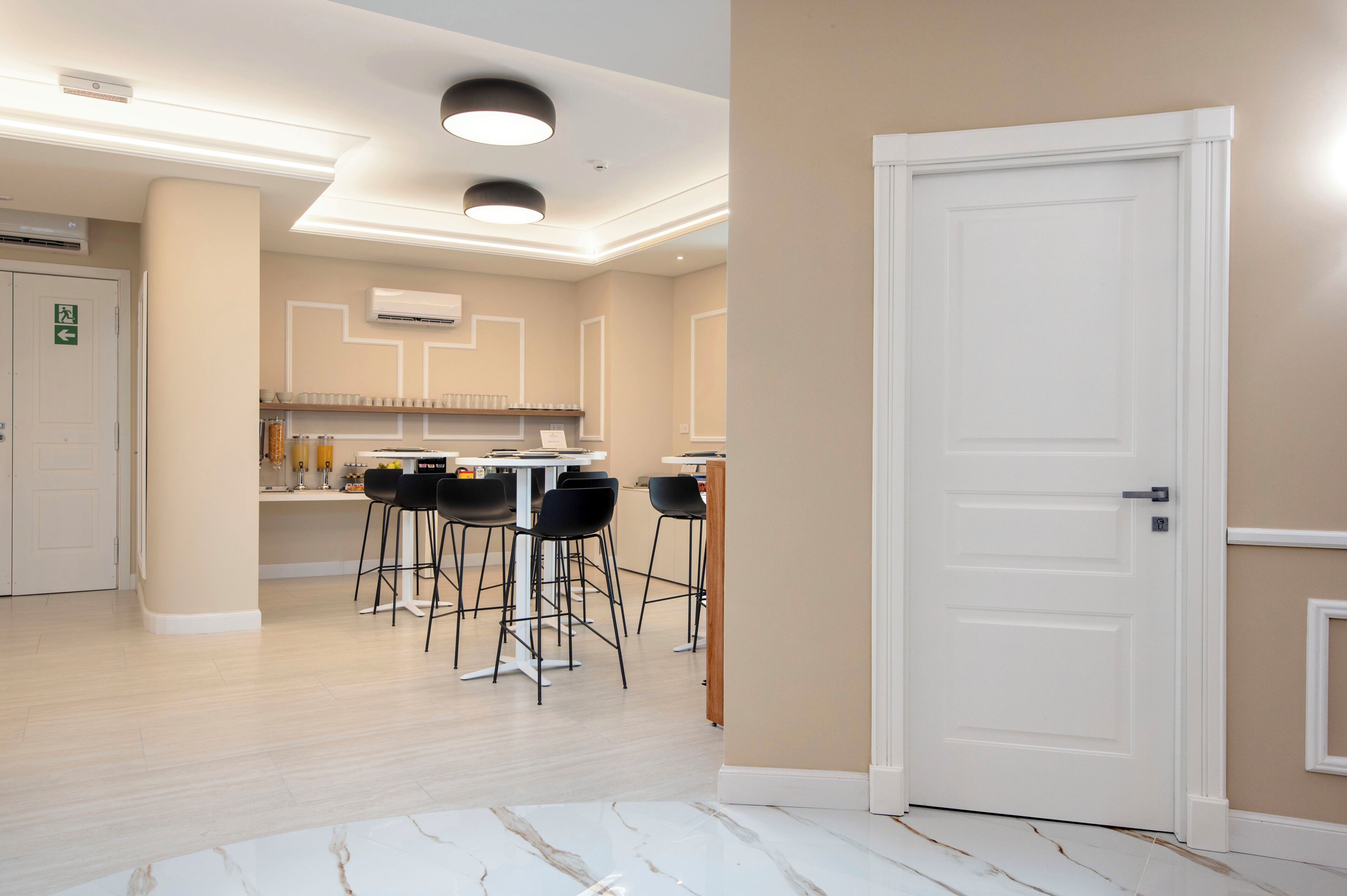 A hotel room with a white door, a small kitchenette, a dining area with four chairs, and a marble floor.