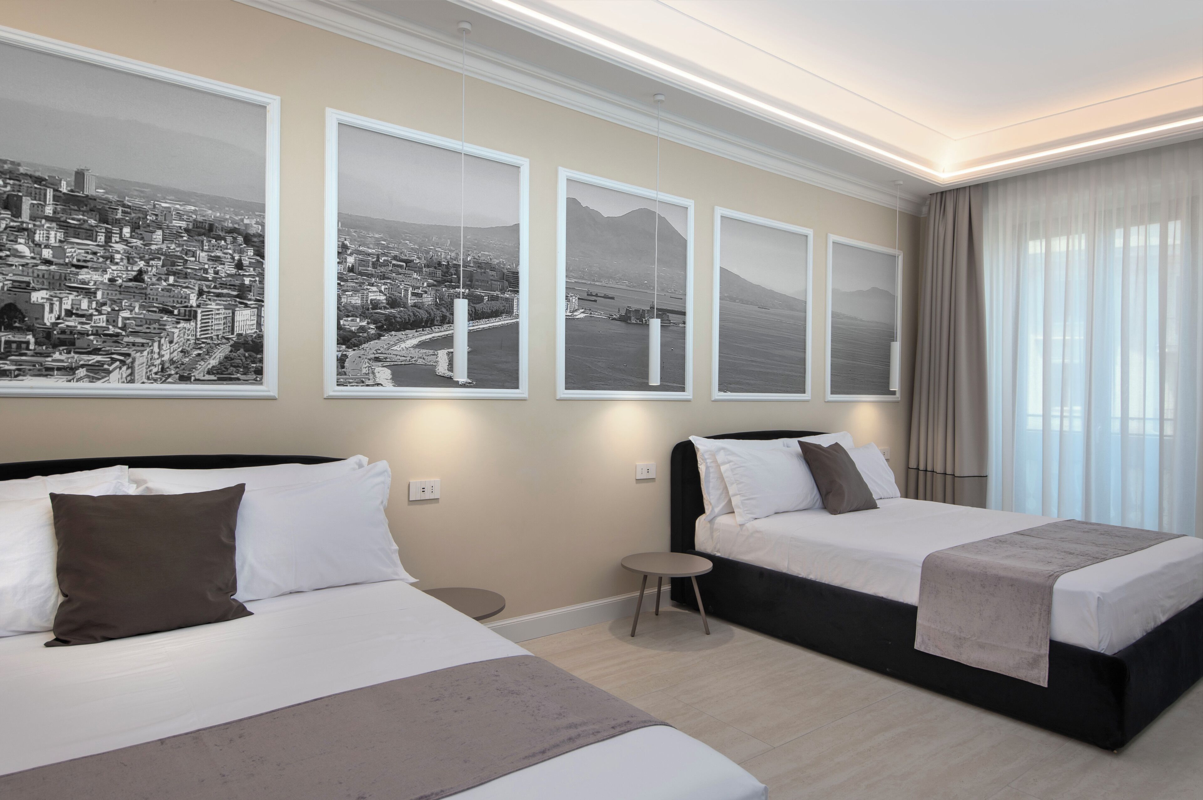 A hotel room with two beds, a small round table, and three framed cityscape photographs on the wall.