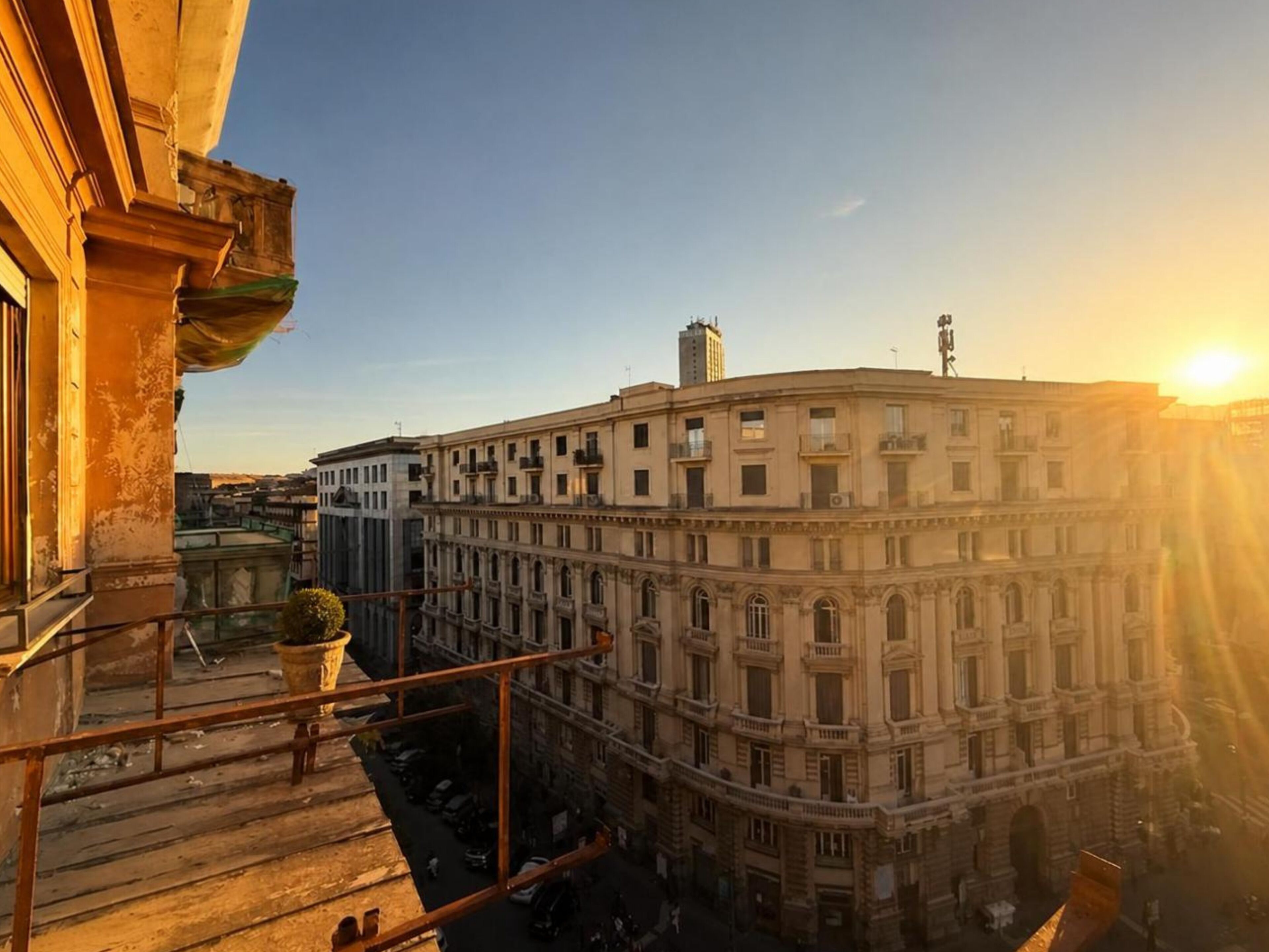 A multi-story building with classical architecture, a balcony with a potted plant, and a clear sky.