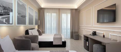 Habitación de hotel con cama, escritorio, silla, taburete pequeño, televisión y ventana con cortinas.