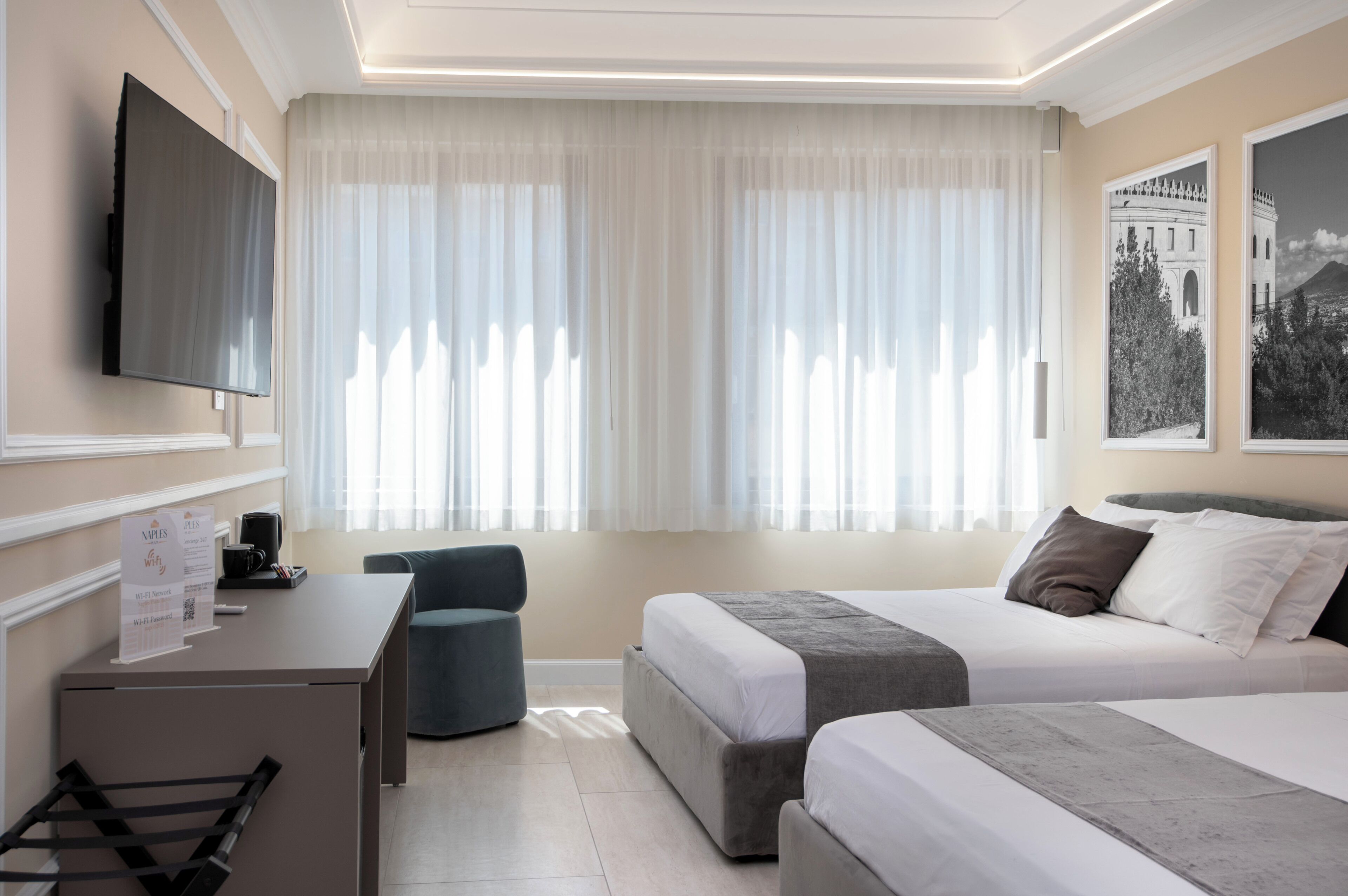 Quarto de hotel com cama, escrivaninha, cadeira, televisão e duas janelas com cortinas finas.