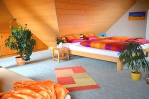 Ein Schlafzimmer mit schrägem Dach, einem Bett mit farbenfroher Bettwäsche, einem kleinen Nachttisch, einer Topfpflanze und einem bunten Teppich.