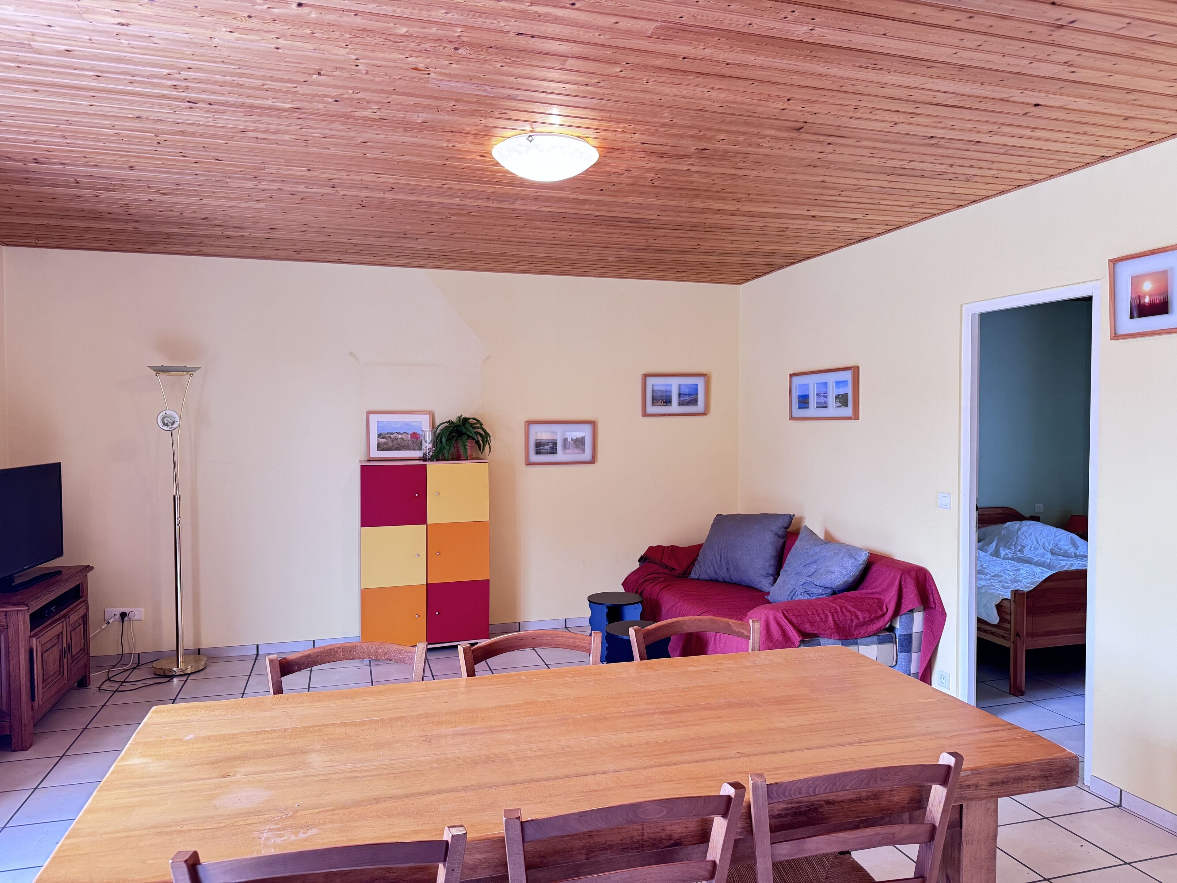 A room with a wooden ceiling, a dining table with chairs, a sofa with cushions, a television, and a cabinet with pictures.
