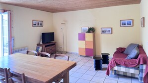 Ein Wohnzimmer mit Holzboden, einem Fernseher, einem Esstisch mit Stühlen, einer Couch mit Decke und einer Vitrine mit Bildern.