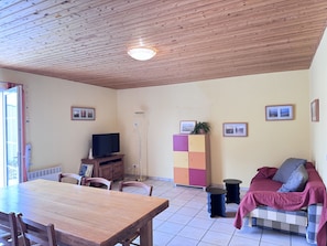 Ein Wohnzimmer mit Holzboden, einem Fernseher, einem Esstisch mit Stühlen, einer Couch mit Decke und einer Vitrine mit Bildern.