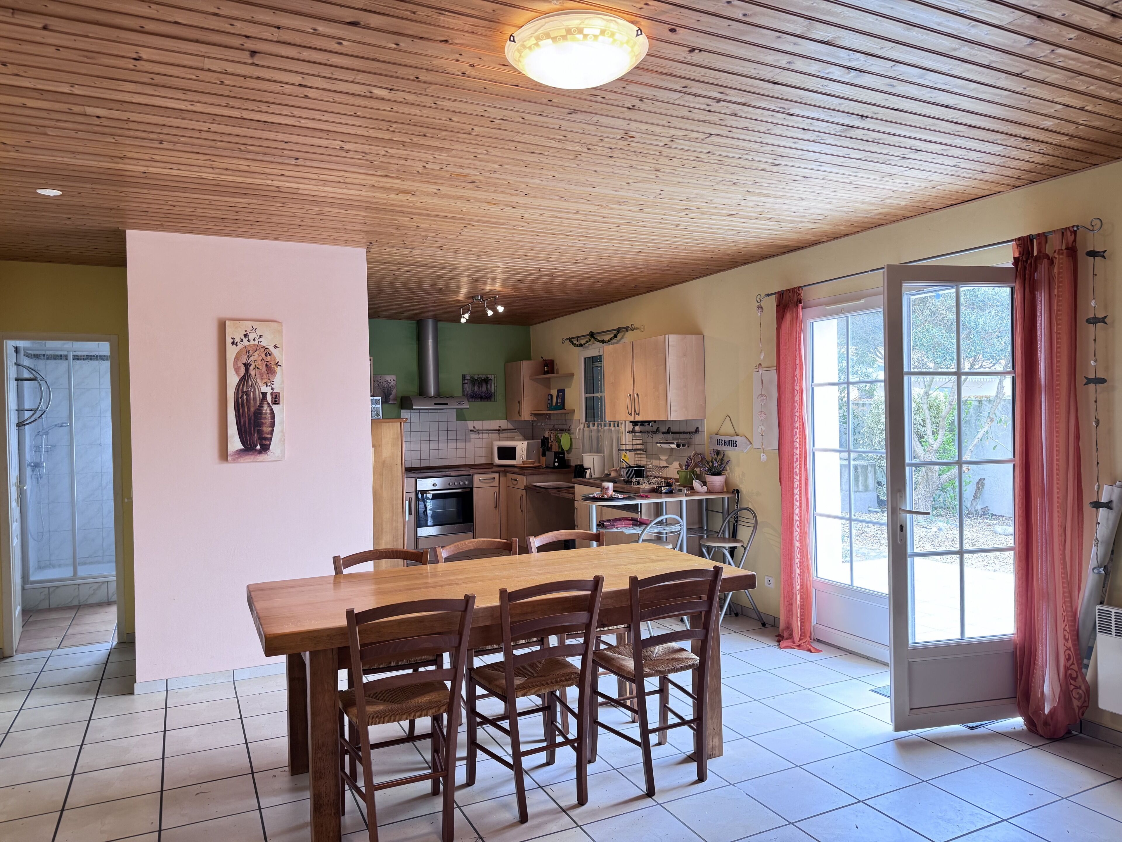 A kitchen with wooden cabinets, a dining table with chairs, a hanging light fixture, and a view through a glass door.