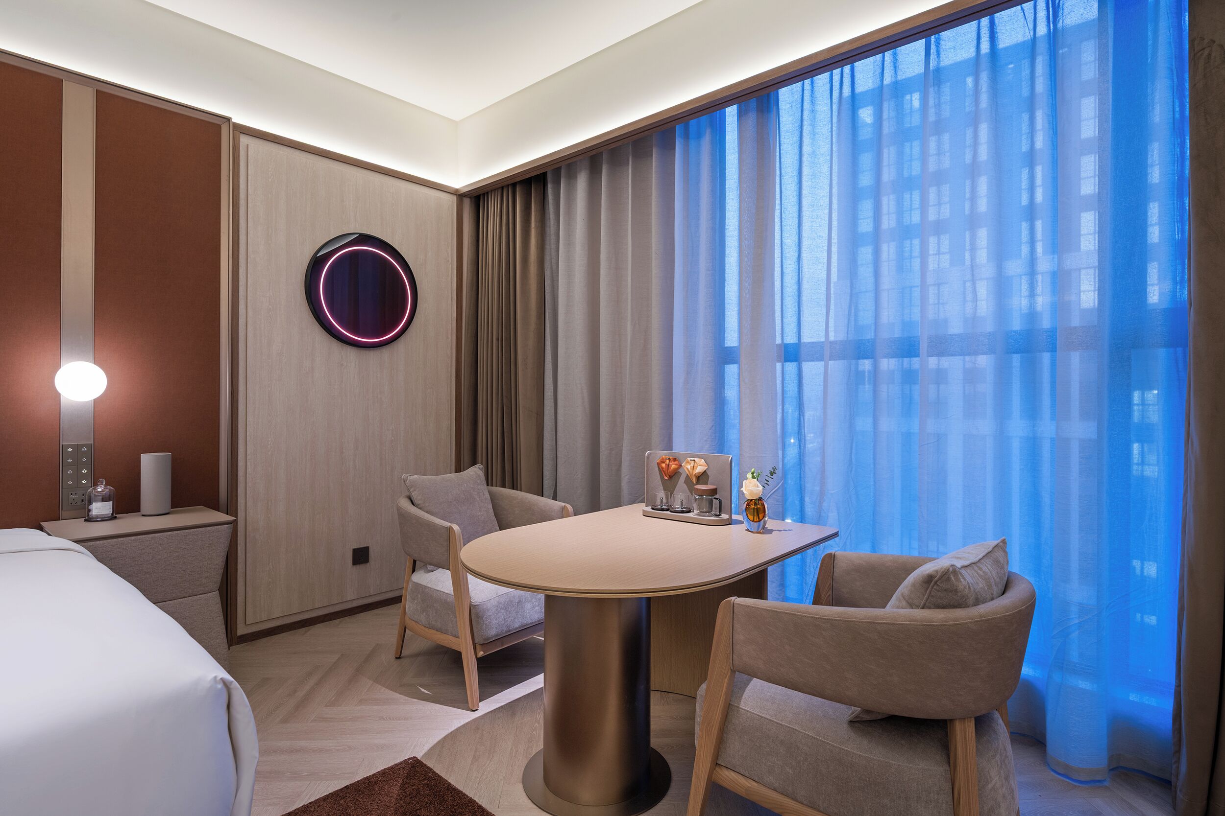 A hotel room with a bed, a round table with chairs, a wall-mounted light, and a view through sheer curtains.