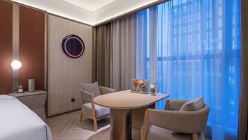 A hotel room with a bed, a round table with chairs, a wall-mounted light, and a view through sheer curtains.