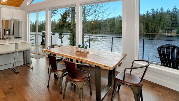 A spacious dining area with a large wooden table and chairs, large windows with a view of a lake and forest. - Modern Lakefront home, 3 bds, 2 baths, dock, gazebo, kayaks, and fishing. (Stanwood)
