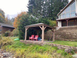 Een overdekt terras met stoelen, een stenen muur en een huis met grote ramen.