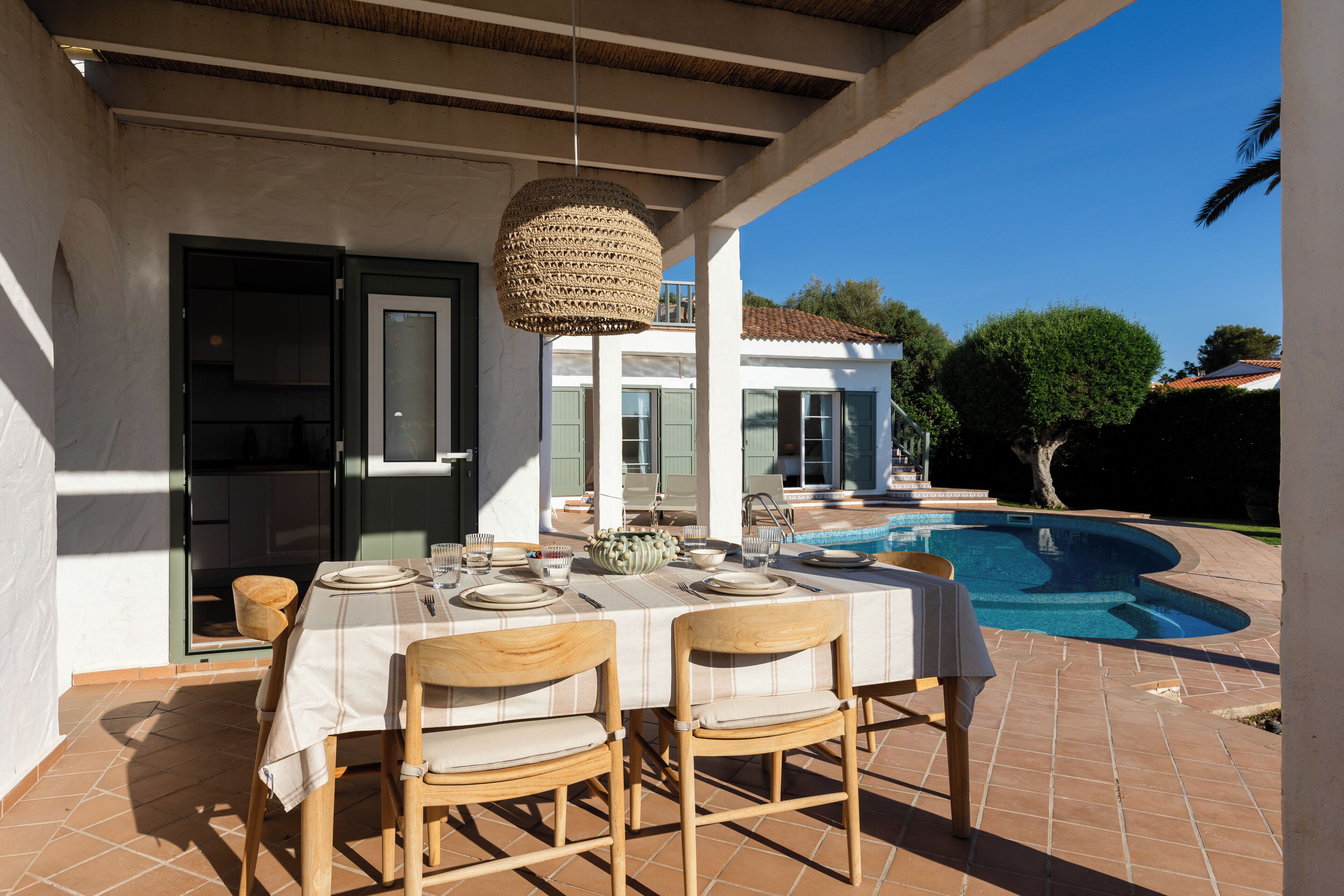 An outdoor dining area with a table set for two, a hanging light fixture, and a swimming pool in the background.